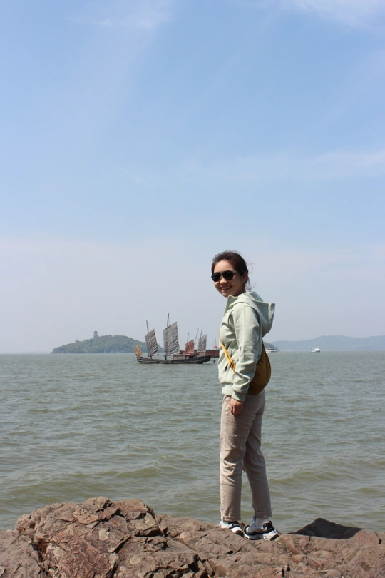 一位站在水体岸边岩石上的女子 远处有一艘船。