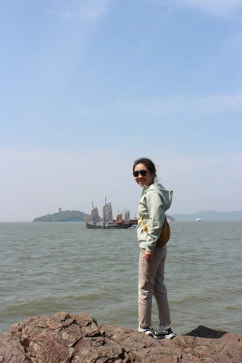 一位站在水体岸边岩石上的女子 远处有一艘船。