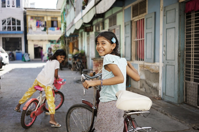 一个小女孩骑着自行车穿过街道两旁有小型建筑的小巷 微笑着。