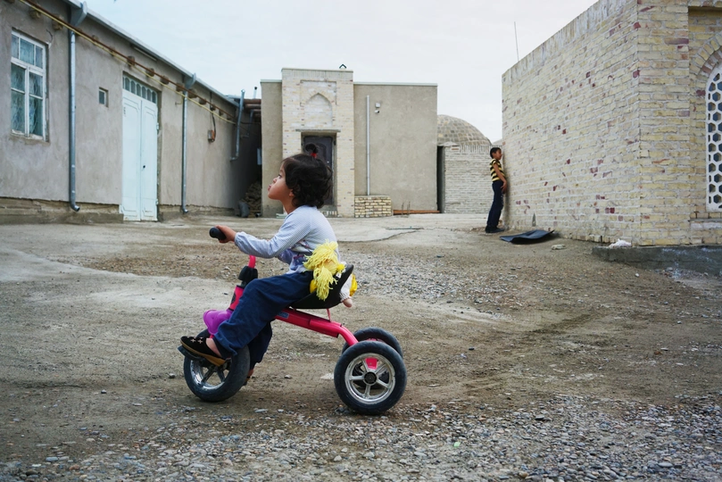 一个小女孩骑着一辆小自行车 车上有只泰迪熊 站在一座破败的建筑前。