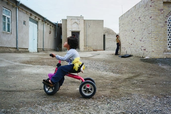 一个小女孩骑着一辆小自行车 车上有只泰迪熊 站在一座破败的建筑前。