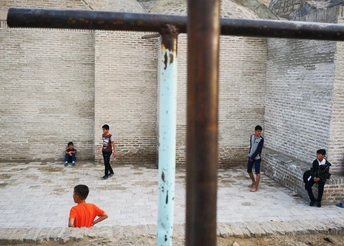 一个穿橙色衣服的人在铁栅栏后面看着小男孩们在街上踢足球。