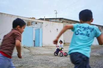 一个小男孩在破败的建筑物后面的小街上踢足球 还有一个小男孩骑着三轮车。