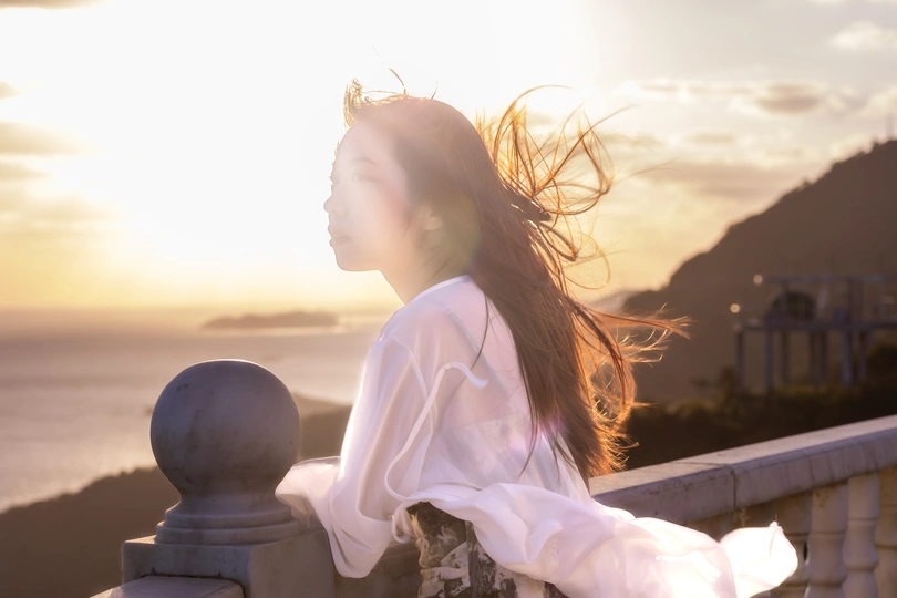 一位年轻女子倚靠在栏杆上 眺望着日落时的海滩 太阳正从栏杆上方露出 她的头发在风中飘动。