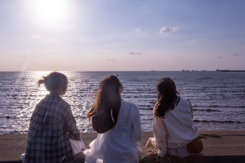 两个年轻女子和一个女孩坐在海滩边的水边 观赏着落日余晖映照在海洋上的美景。