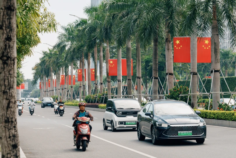 一条街道 两边种着棕榈树 停满了汽车 还有骑摩托车和滑板车的人们。