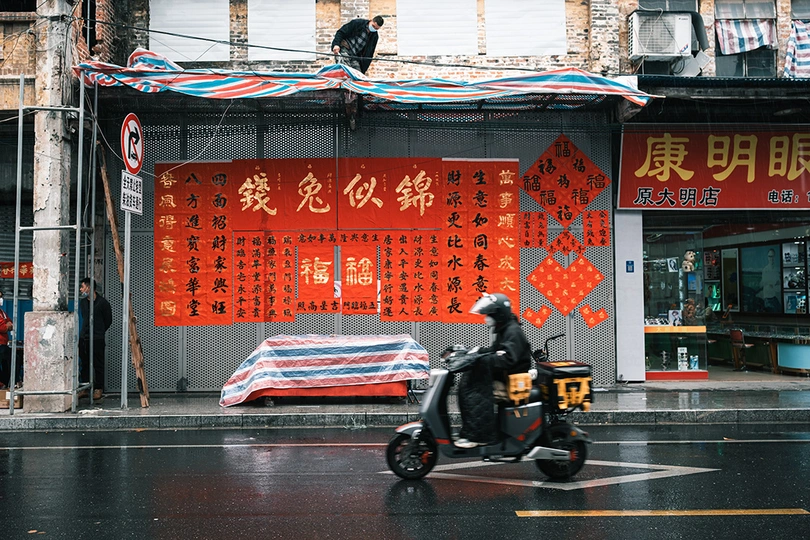一个摩托车手在雨中驶过一所建筑 该建筑上写有中文。还有一个男人在雨中骑着滑板车。
