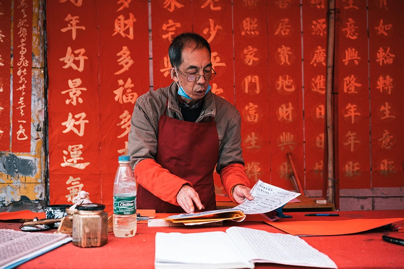 一个戴红围裙的男子坐在桌子上 桌子上放着笔记本和剪刀。