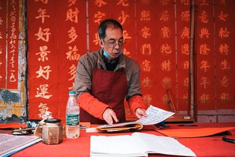 一个戴红围裙的男子坐在桌子上 桌子上放着笔记本和剪刀。