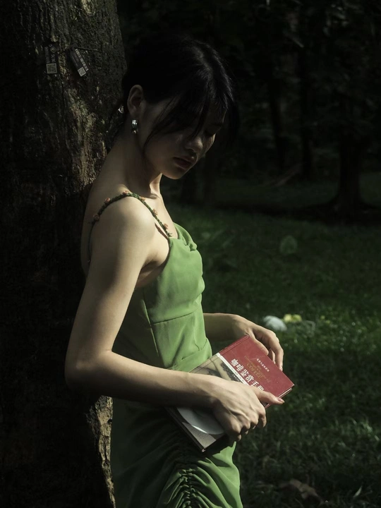 一位穿着绿色连衣裙的年轻女子 手里拿着一本书 靠在一棵树旁。