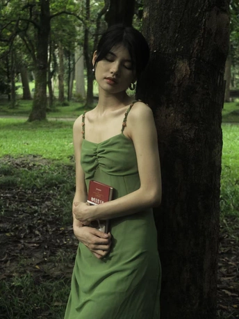 一位穿着绿色连衣裙的年轻女子站在森林中的一棵树旁边 手里拿着一本书。