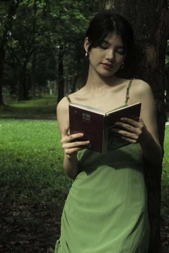 一位穿着绿色连衣裙的年轻女子正在一棵树旁的公园里阅读一本书。
