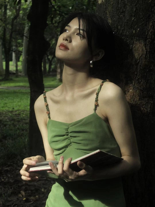 一位穿着绿色连衣裙的年轻女子手里拿着一本书站在一棵树旁边
