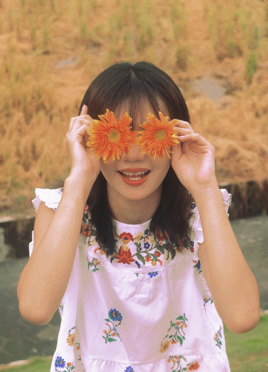 一个小女孩穿着花裙子 用手捂住脸 用花遮住眼睛。
