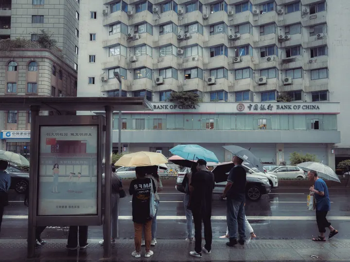 一些人站在公交车站 在雨中 前面有一些建筑 他们手里拿着伞。