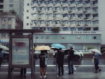 一些人站在公交车站 在雨中 前面有一些建筑 他们手里拿着伞。