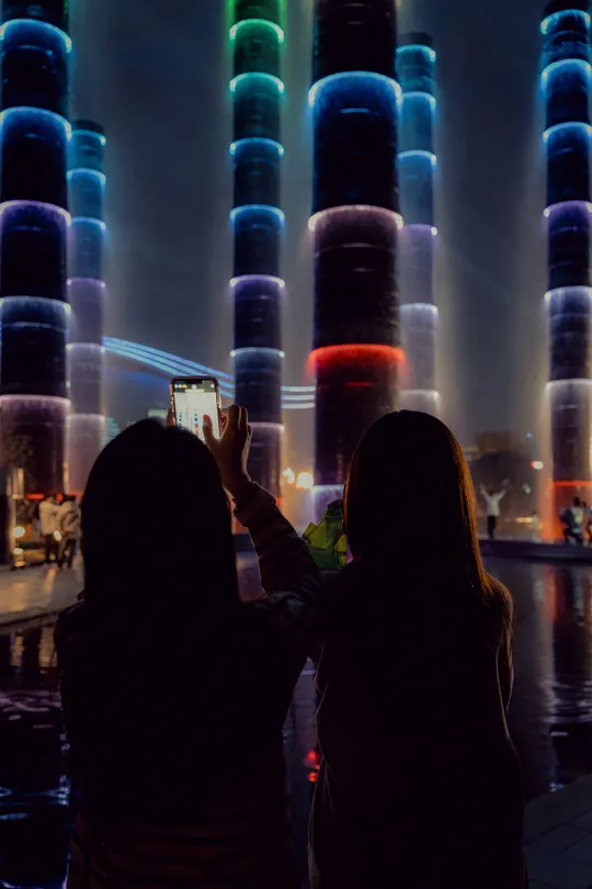 一位女性用手机拍摄夜晚一座高塔上的灯光 周围有人们站立。