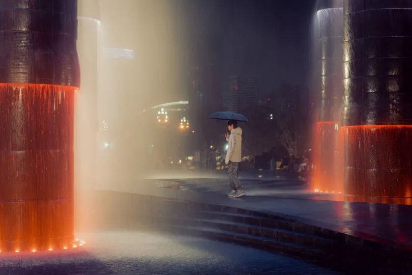 一位女士在夜晚穿过城市 手里拿着一把雨伞 经过一个红色的喷泉。