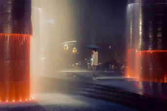 一位女士在夜晚穿过城市 手里拿着一把雨伞 经过一个红色的喷泉。