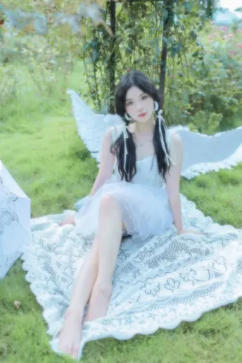 一个穿着白色连衣裙有翅膀的女孩坐在草地上的一块毛毯上