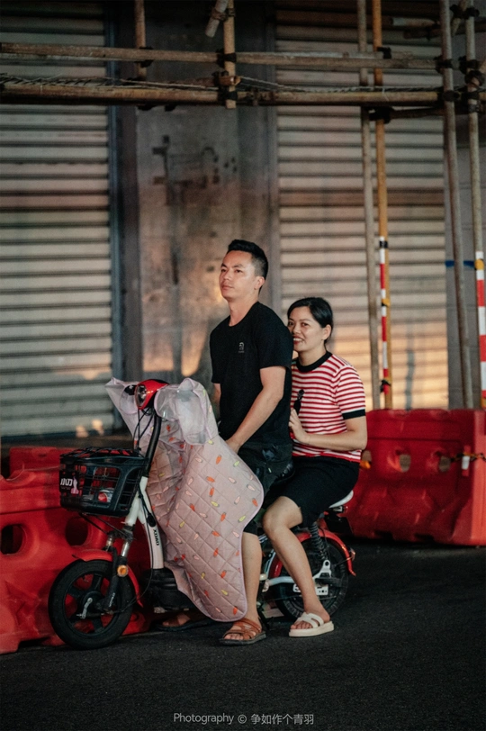 一个年轻男子和女子坐在红色滑板上在街上 而一个年轻男子在他们身后骑自行车。