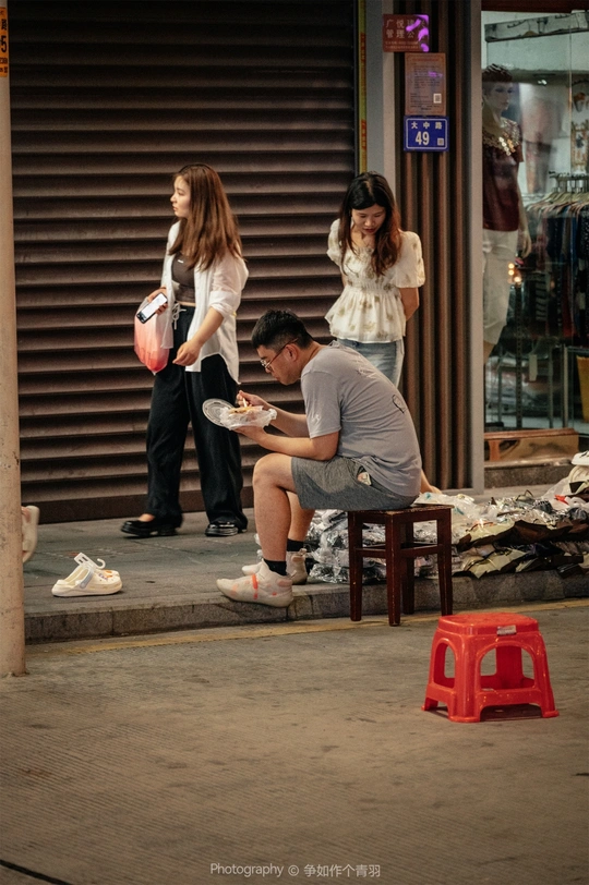 一个年轻男子坐在小板凳上 从碗里吃东西 而一个妇女站在他身后的人行道上。