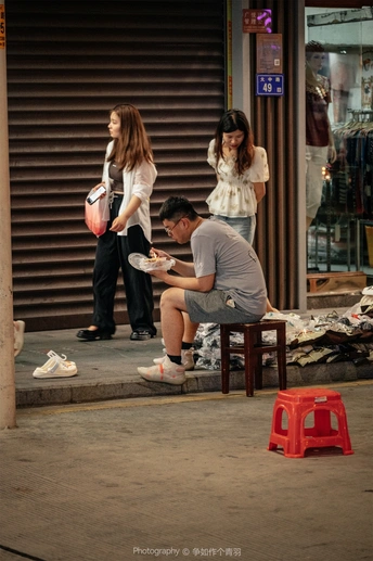 一个年轻男子坐在小板凳上 从碗里吃东西 而一个妇女站在他身后的人行道上。