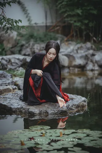 一个穿着黑色和红色衣服的女孩坐在池塘边一块岩石上