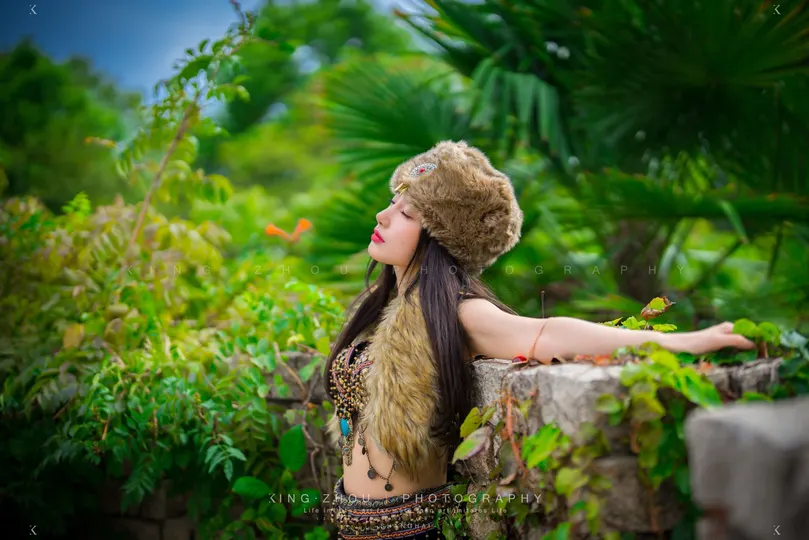 一个长发女子戴着帽子站在丛林中的石头墙上