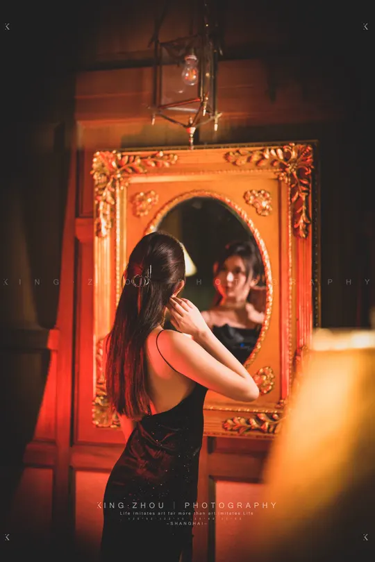 一位穿着黑色连衣裙的年轻女子站在一个红色的房间里 站在镜子前。