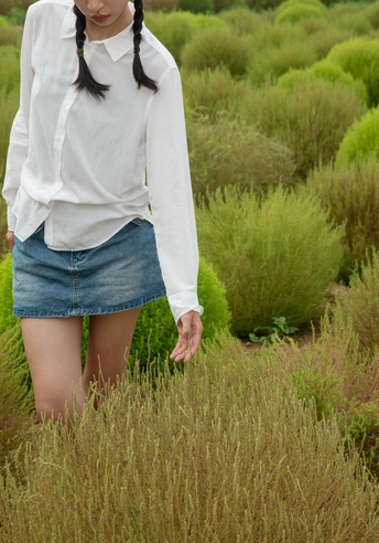 一位穿着白衬衫和裙子的年轻女子穿过高草和灌木丛。