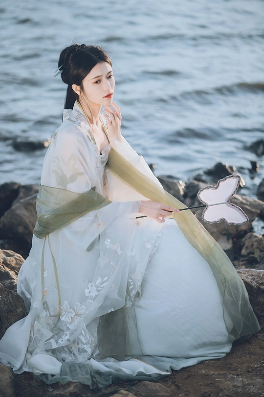 一位穿着白色连衣裙的美女坐在水边的一块岩石上