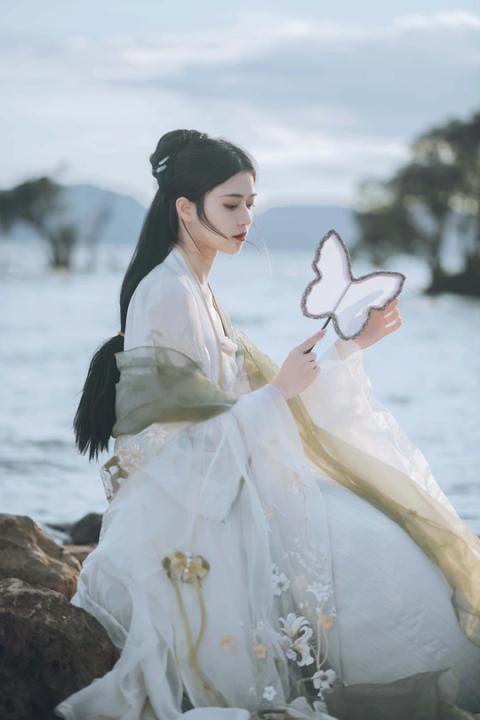 一位穿着传统裙子的美丽女孩 蝴蝶停在岩石上 岩石靠近水边。