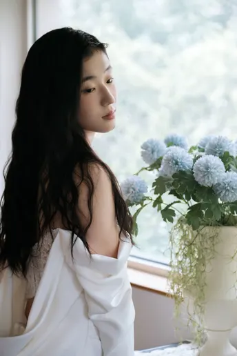 一个长发着白裙子的年轻女子坐在窗边 旁边放着一束花。