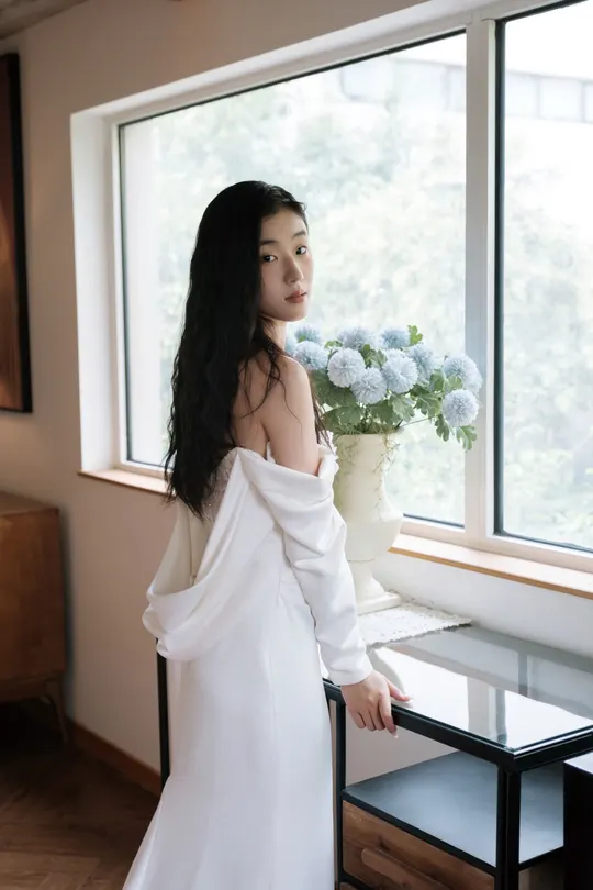 一位穿着长白色连衣裙的女人站在窗户前 手里拿着一束花。