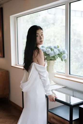 一位穿着长白色连衣裙的女人站在窗户前 手里拿着一束花。