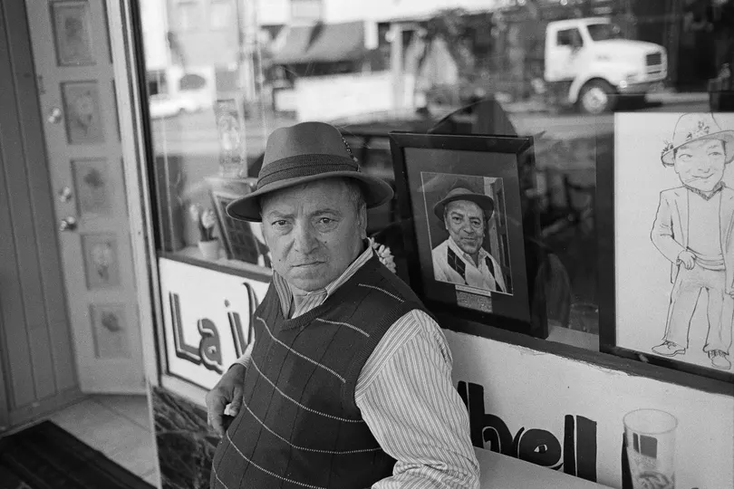 一张黑白照片 一位老人戴着帽子站在窗前 凝视着一幅画。