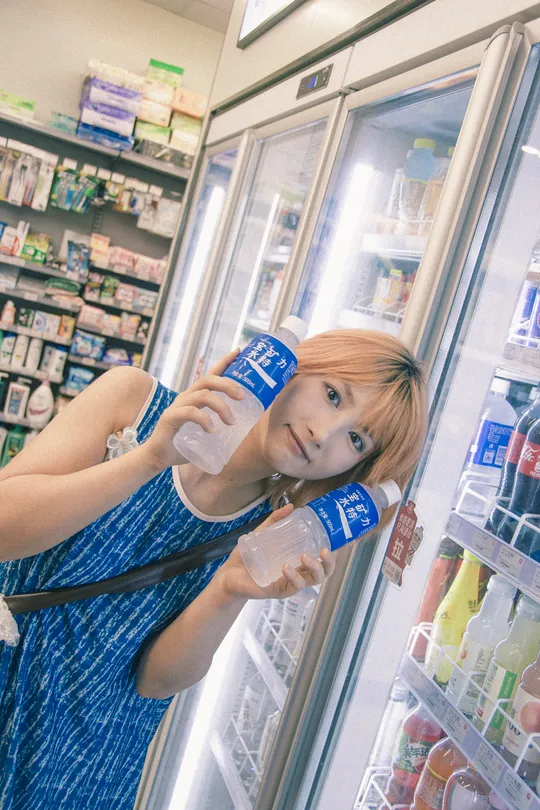 一位年轻女子手持一瓶水和一个杯子站在一家商店的冰箱前面。