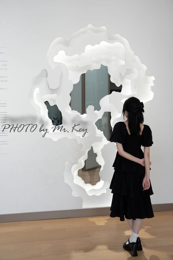 一位穿着黑色连衣裙的女人正在看墙上有一朵白色云的艺术品。