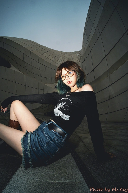 一位戴眼镜的绿发年轻女子坐在建筑物前的一堵墙上