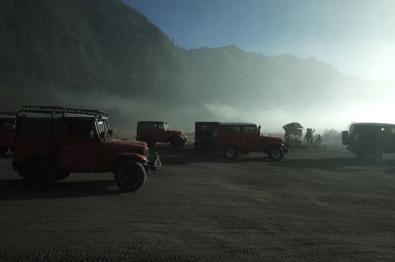 几辆车停在一个停车场里 一辆红色的吉普车在阳光下在山脉前排队行驶。