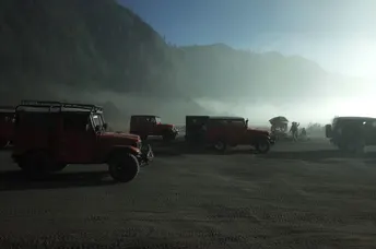 几辆车停在一个停车场里 一辆红色的吉普车在阳光下在山脉前排队行驶。