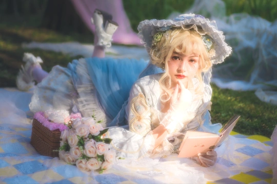 一位穿着白色连衣裙的年轻女子正在一块蓝色和白色格子毛毯上阅读一本书。
