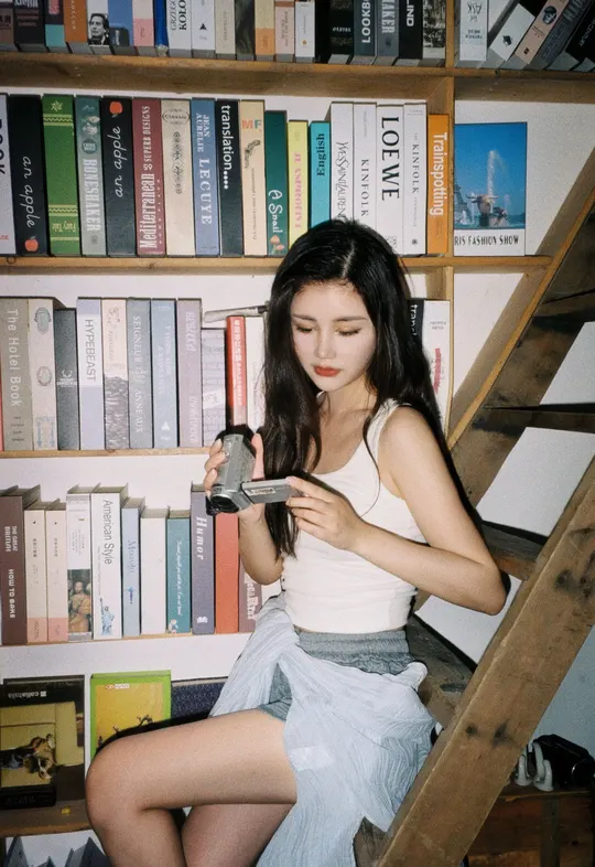 一个穿着白裙子的年轻女子抱着相机坐在书架上