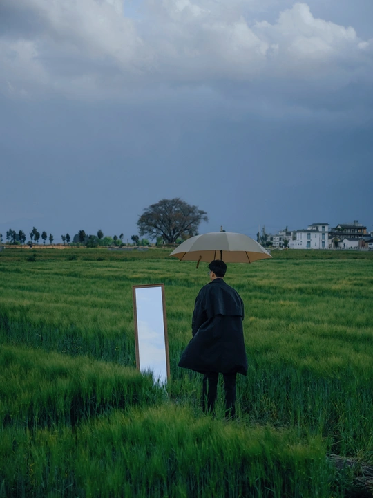 一个人站在草地上 手里拿着一把雨伞 旁边有一个镜子。