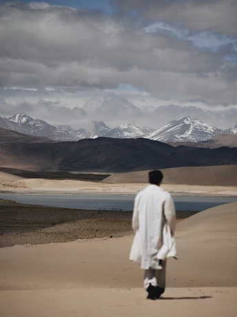 在沙漠中 一个穿着白色长袍的人在湖边走沙子 背景是山脉。