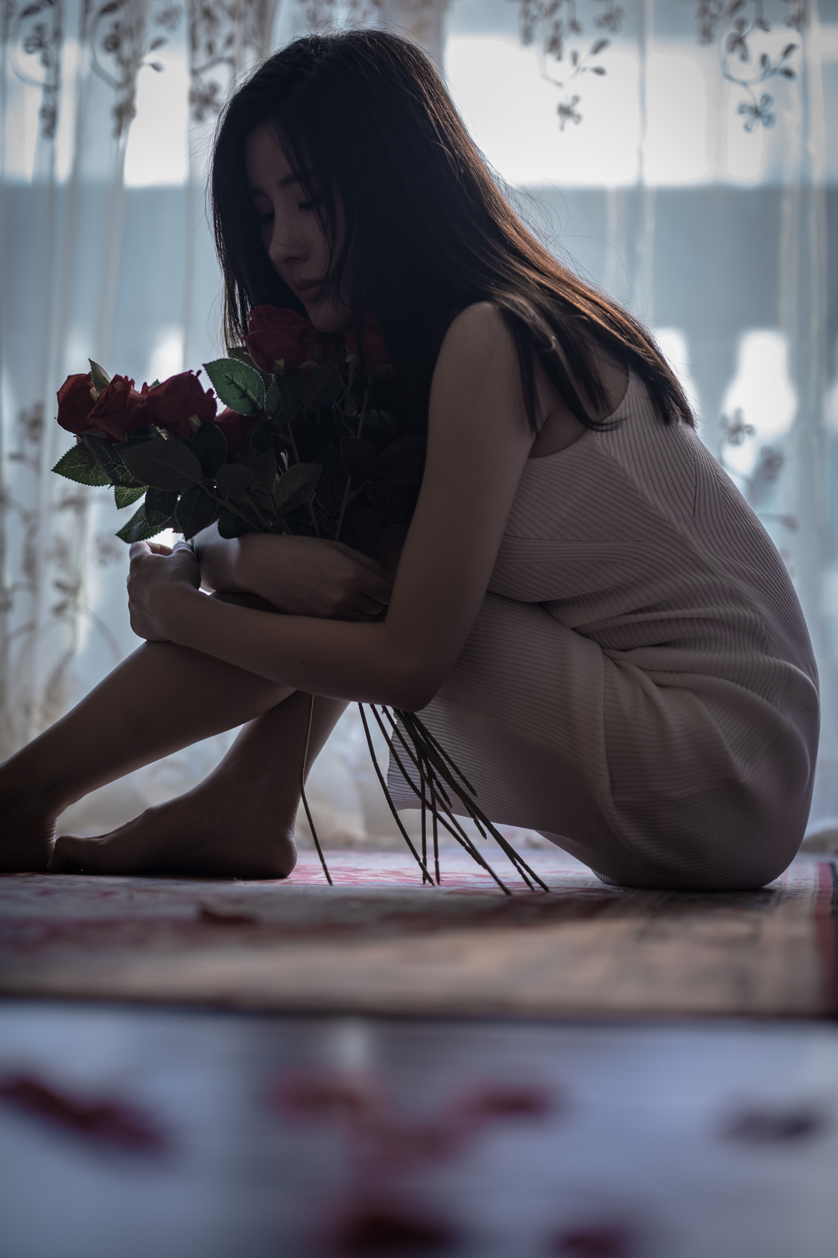 一位年轻女子拿着一束红玫瑰坐在窗前的地板上