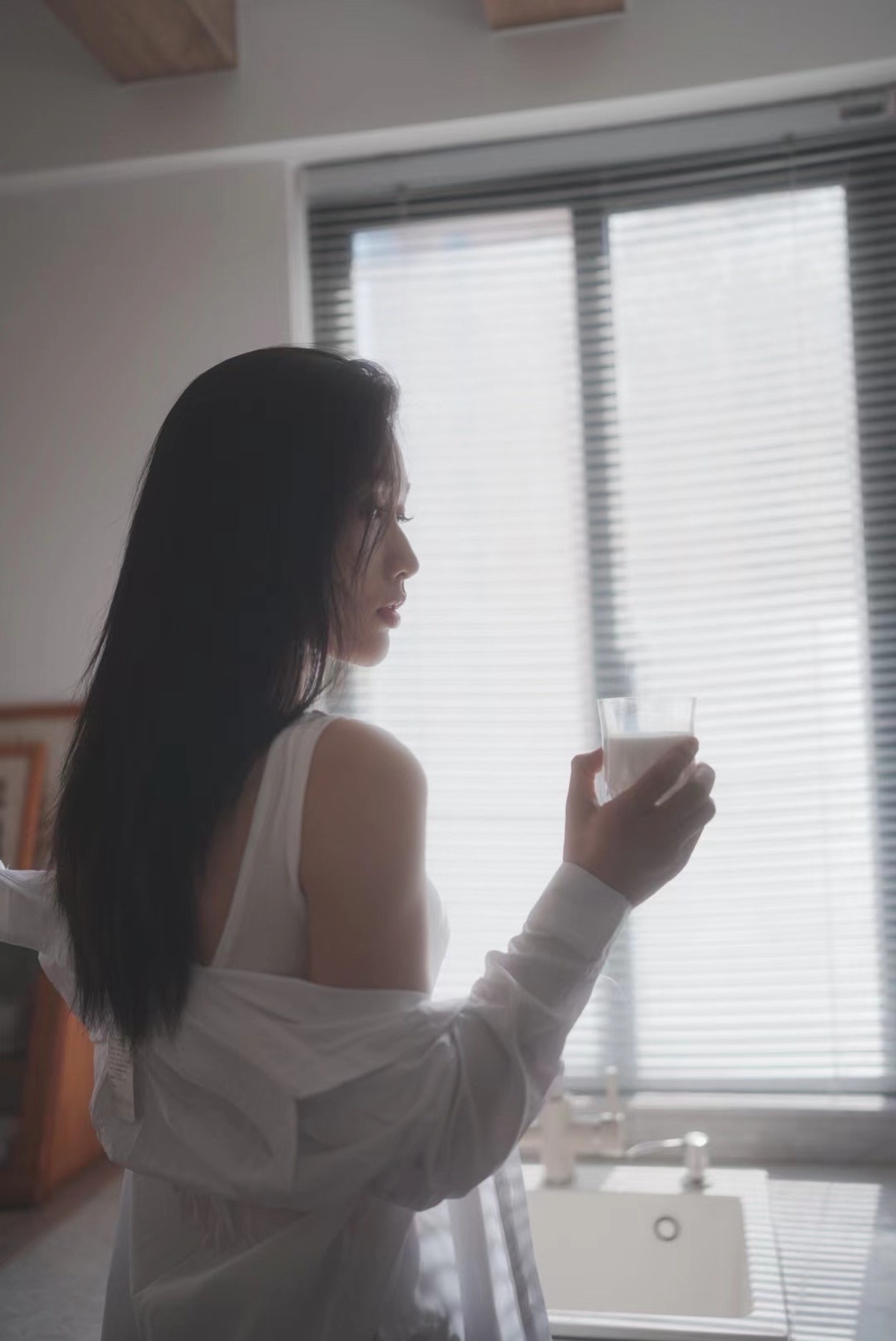 一位年轻女子手持一杯牛奶和一杯水站在窗户前