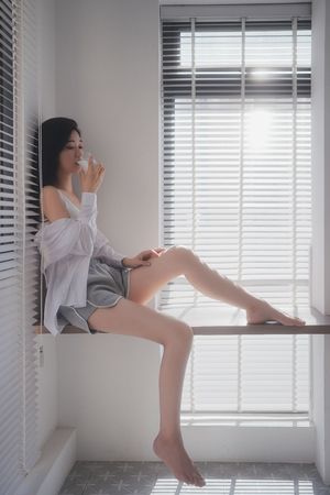 一个年轻女子坐在窗台上 双腿交叉 手里拿着一杯咖啡或茶。