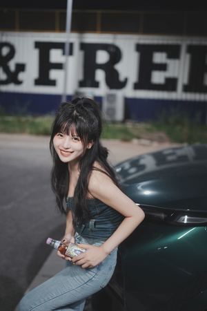 一位年轻女子倚靠在一辆黑色跑车的车盖上 手里拿着一瓶饮料 正在摆姿势拍照。